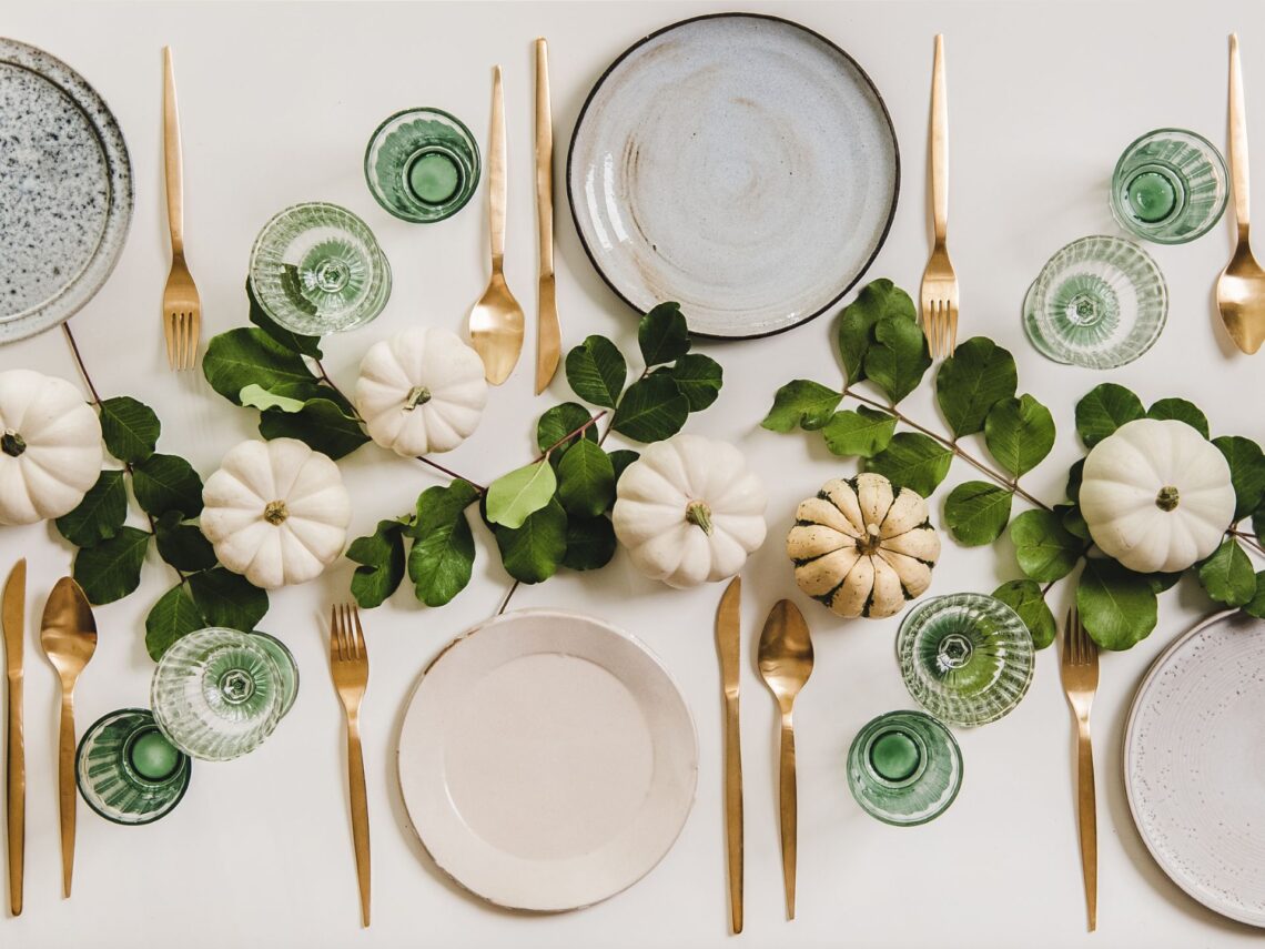 Table settings with plates, gold silverware, emerald green glasses, and white pumpkins.