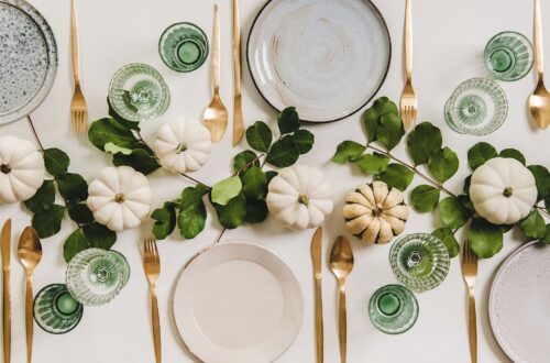 Table settings with plates, gold silverware, emerald green glasses, and white pumpkins.