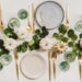 Table settings with plates, gold silverware, emerald green glasses, and white pumpkins.