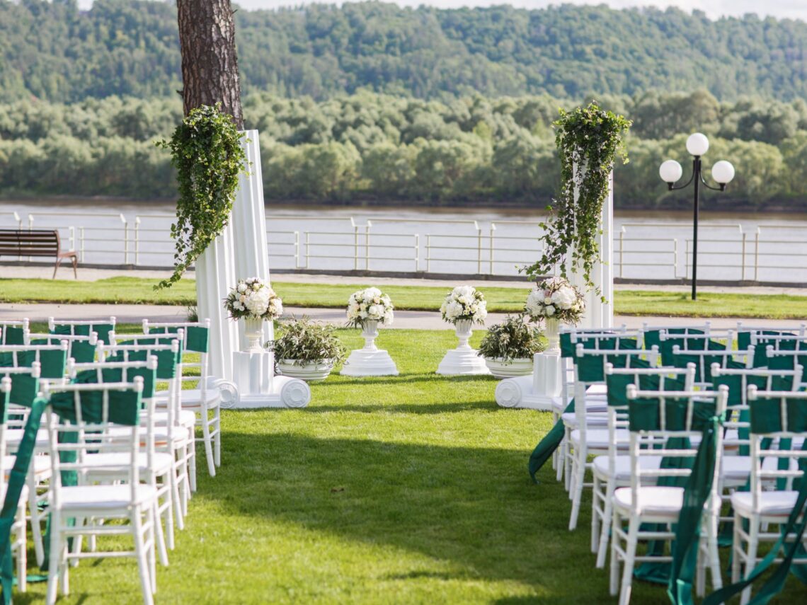 An outdoor wedding venue with white chairs that have emerald green sashes.